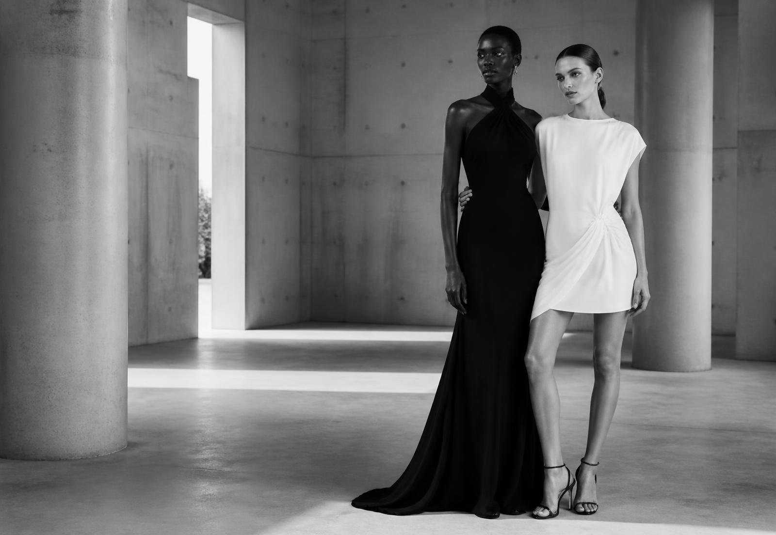 Two models in a black floor-length gown and a white mini dress in a sunlit gallery